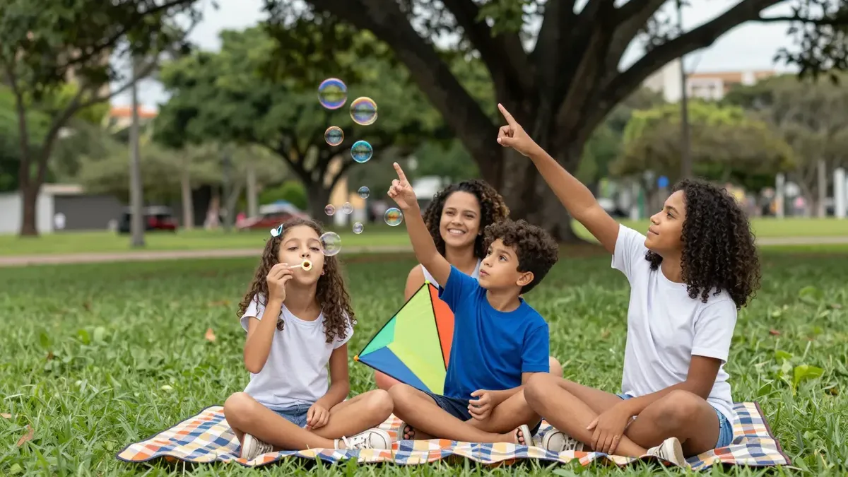 Três irmãos num piquenique sobre uma toalha xadrez no gramado de um parque arborizado, a menor fazendo bolhas de sabão, o menino com uma pipa colorida no colo e a mais velha apontando para o céu, com a mãe sorrindo atrás