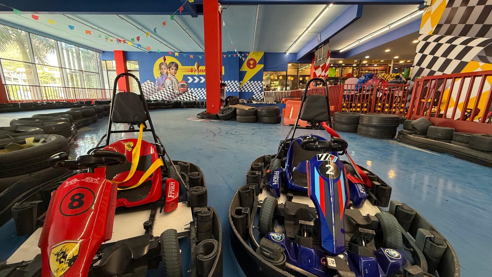 Dois karts elétricos infantis, um vermelho número 8 e um azul número 2, na pista indoor do eKarts com proteção de pneus ao redor