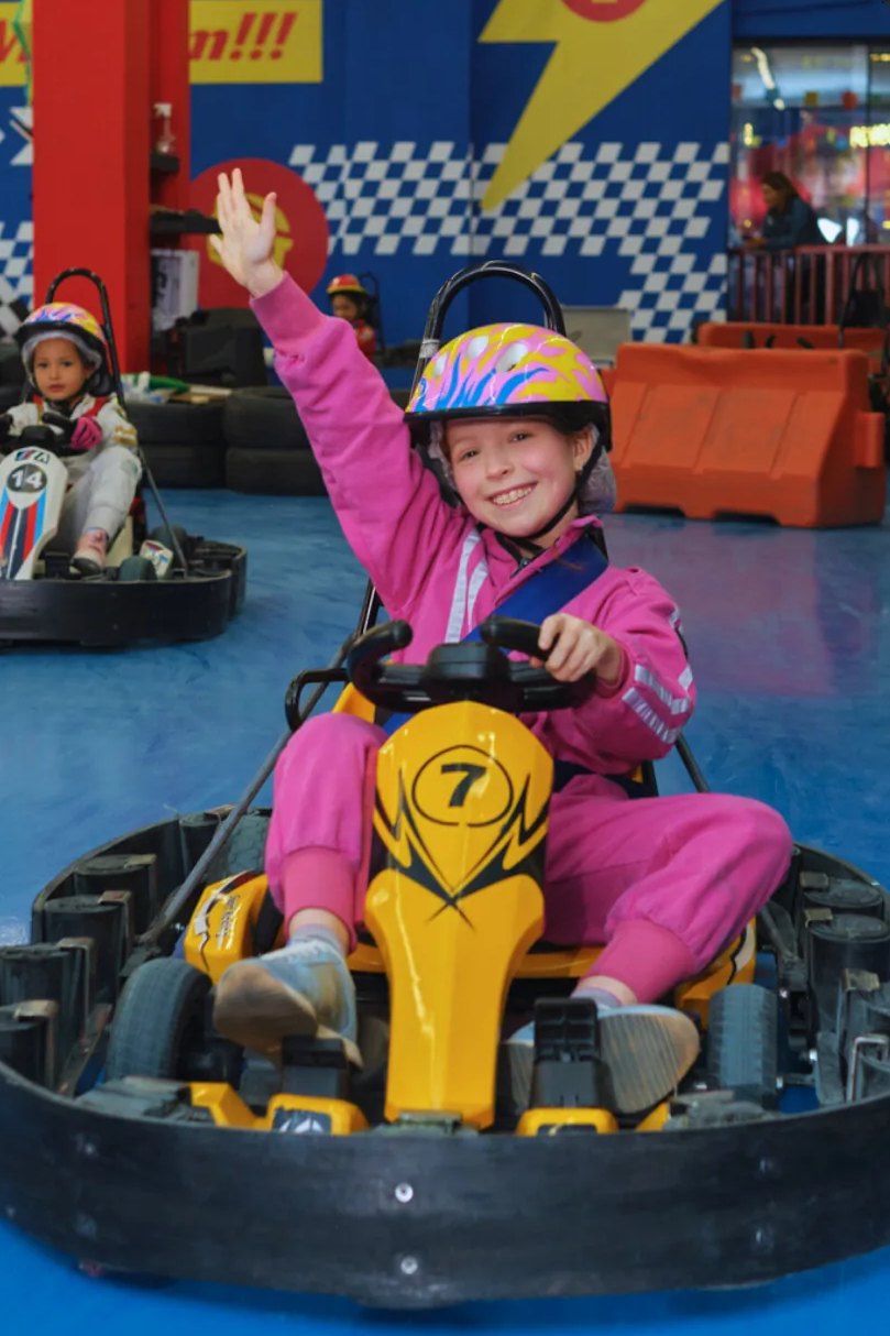 Menina sorrindo de orelha a orelha e acenando enquanto pilota o kart elétrico na pista